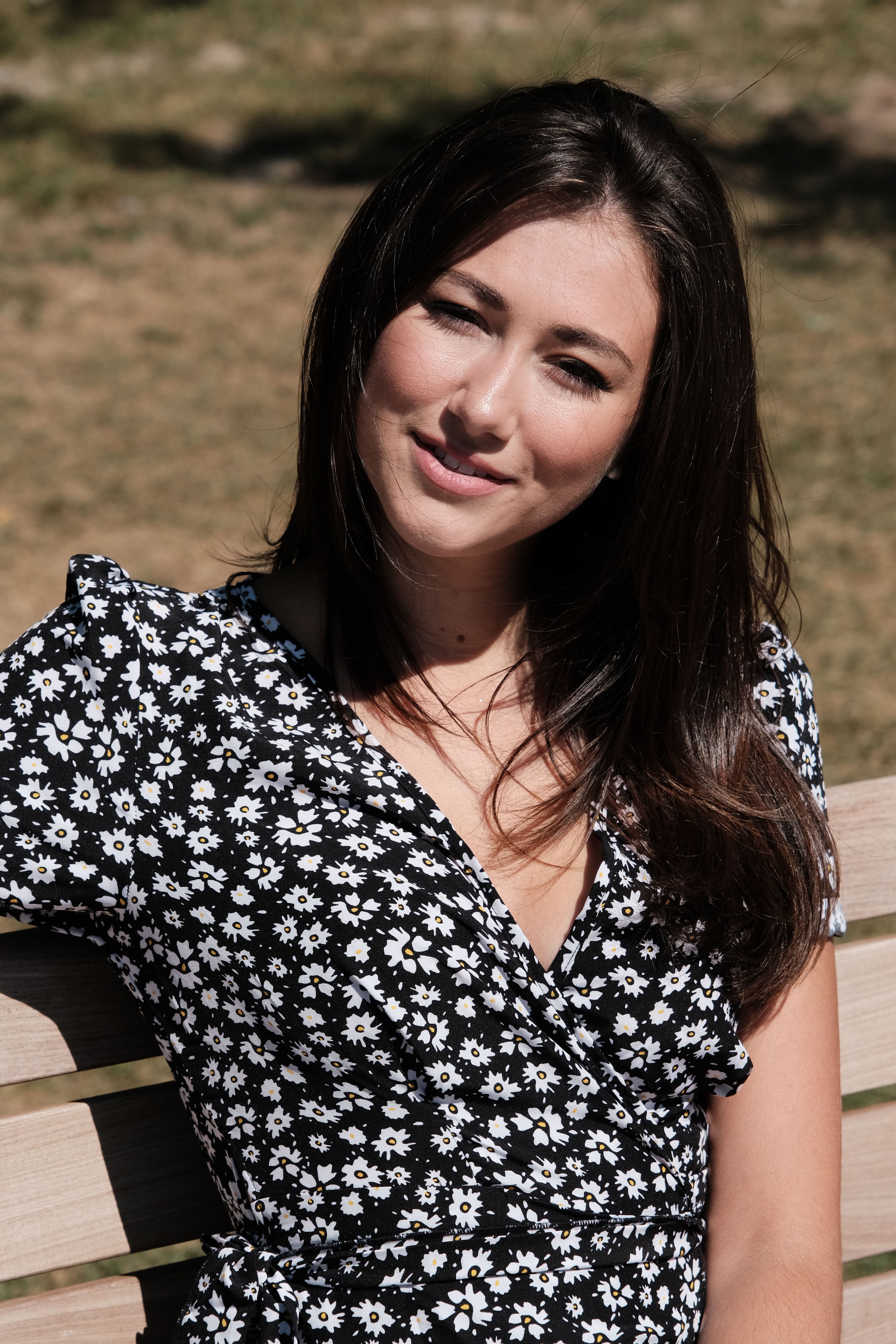 Cheerful woman in a floral dress smiles while sitting on a bench in a sunny park.