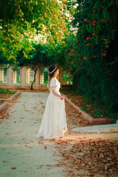 A woman in a stylish white dress walks gracefully in a lush garden in Adana, Turkey, under vibrant foliage.