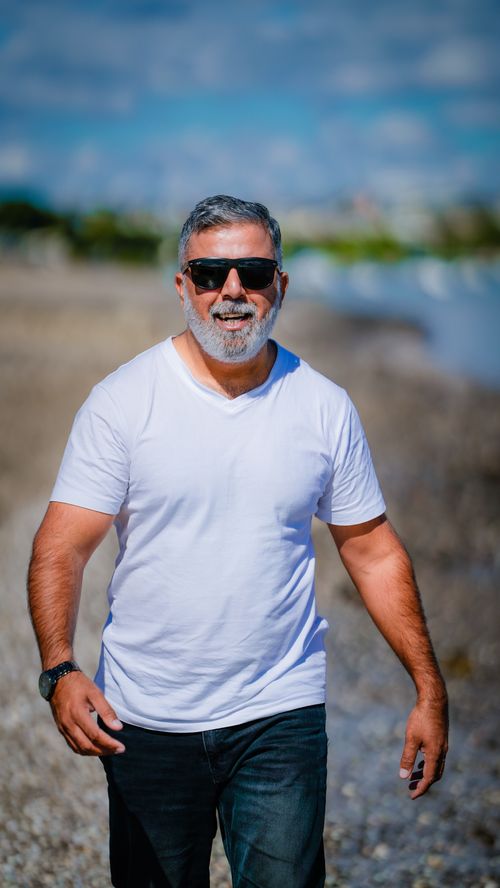 Free Middle-aged man with beard and sunglasses smiling while walking on Auckland beach. Stock Photo