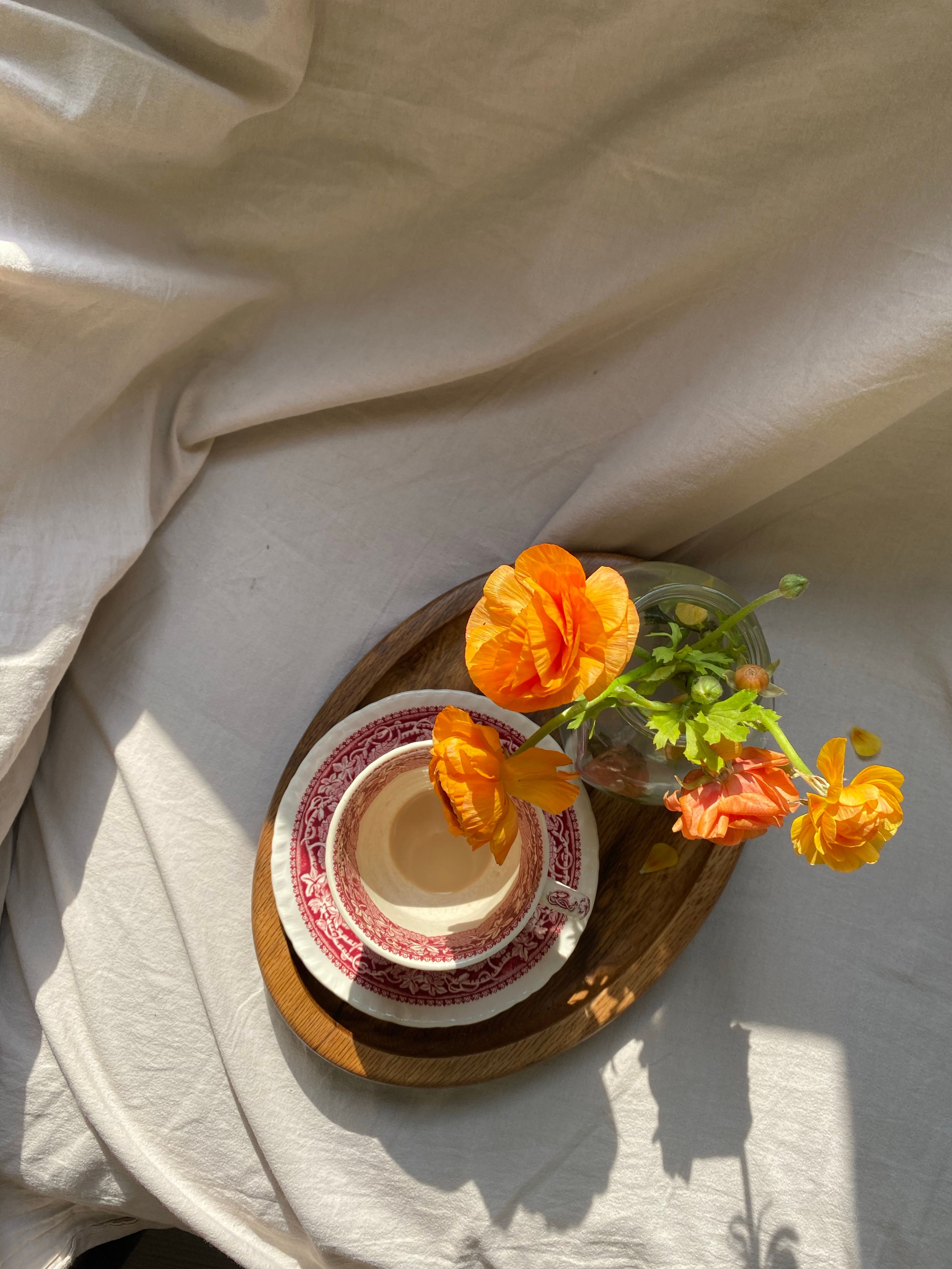 A wooden tray with a teacup and flowers in sunlight on fabric background.