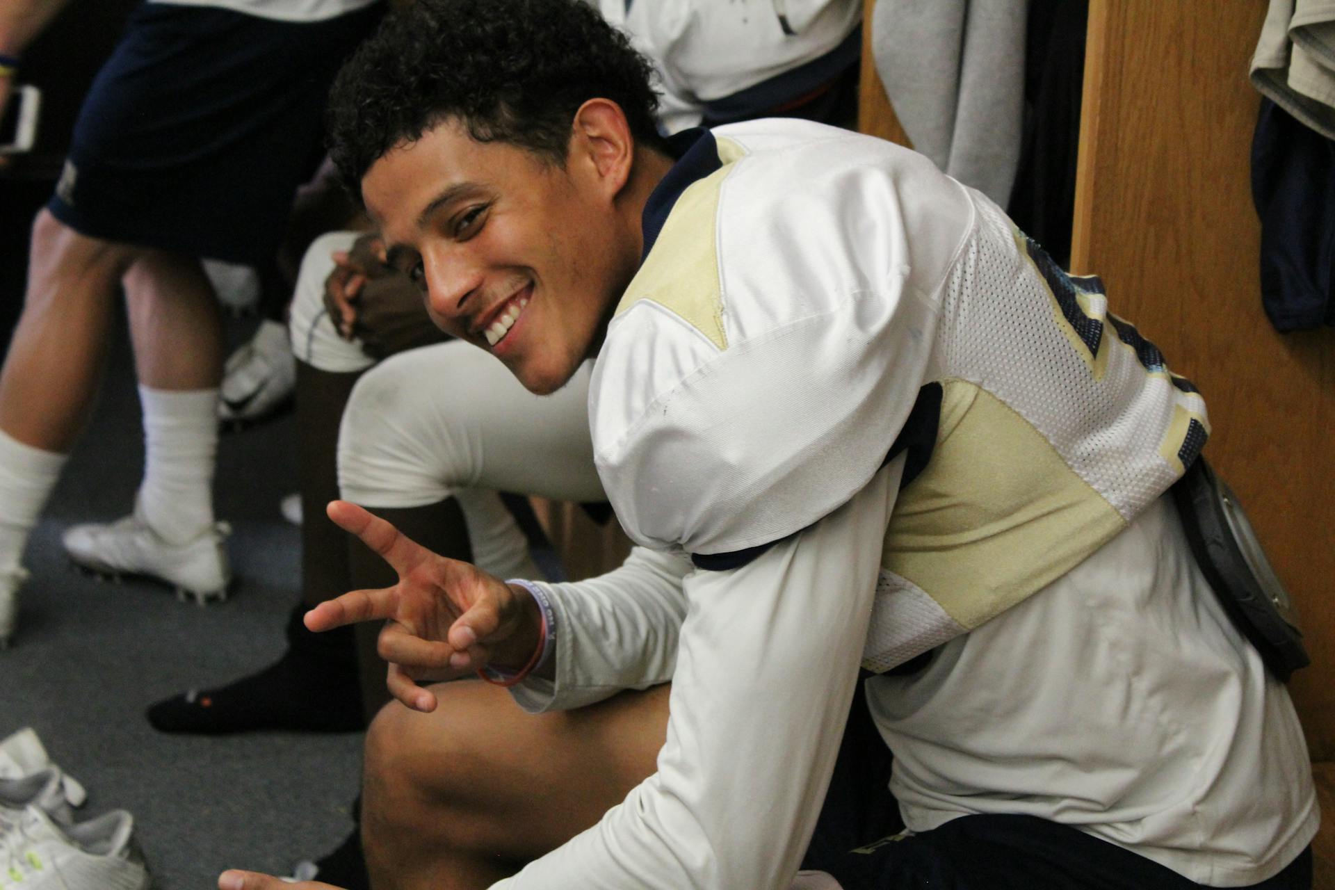 Young American football player smiling and gesturing peace sign inside a team locker room.