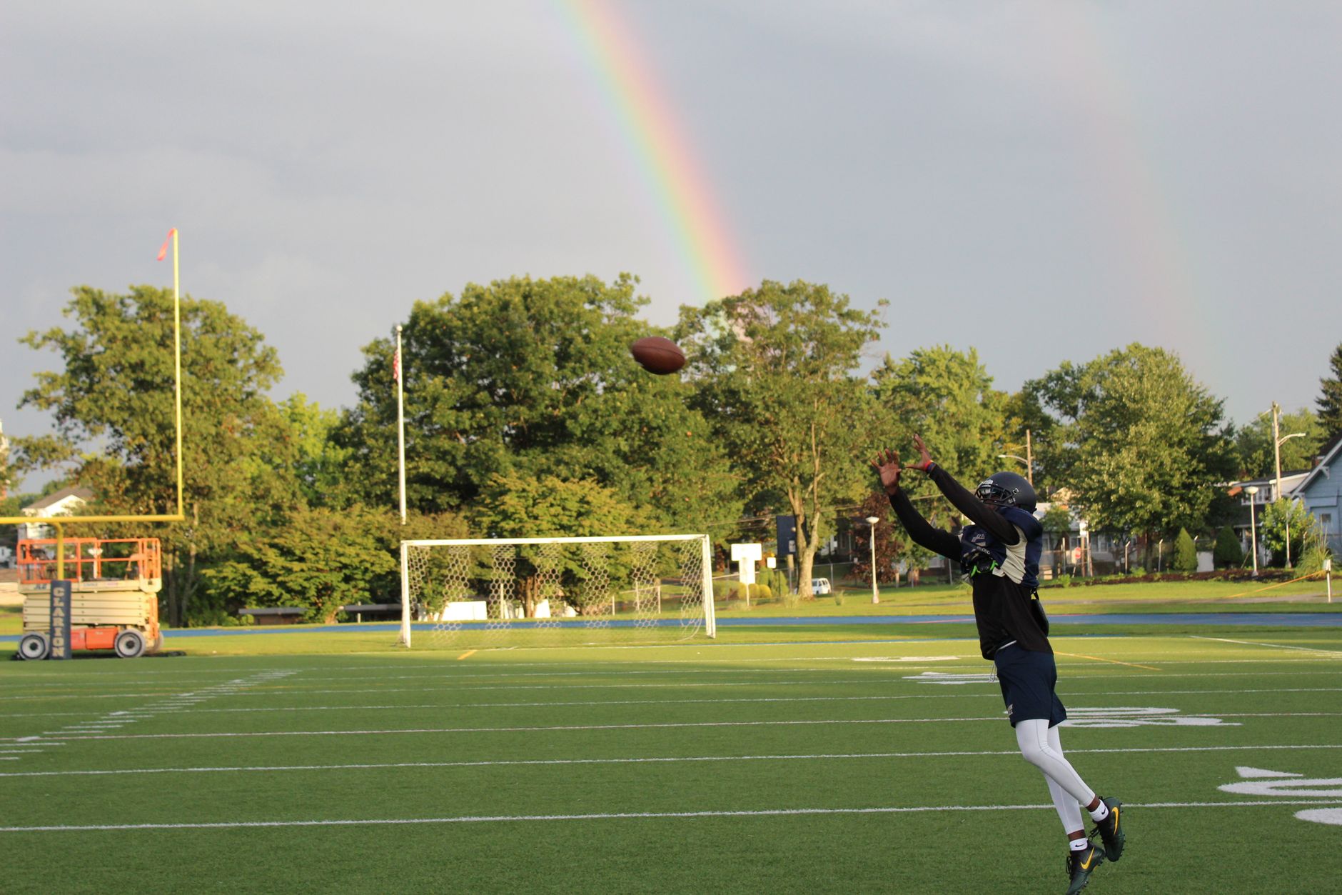 Football player leaps to catch a ball with a vibrant rainbow backdrop on a sunny field.