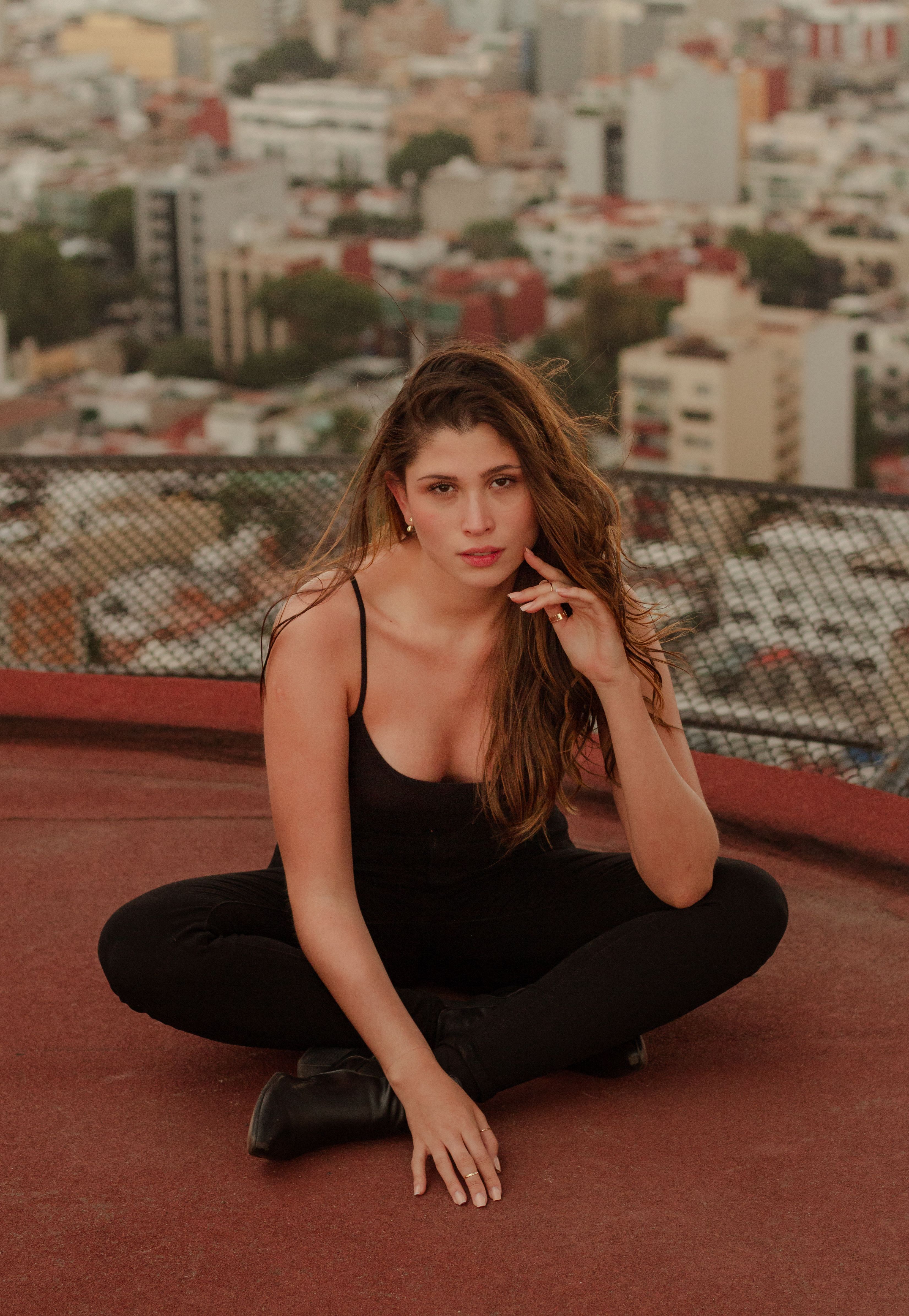 A young woman with long hair poses casually on a rooftop with a vibrant cityscape background.