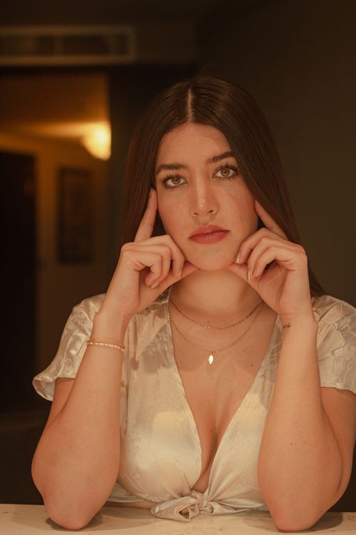 Free Woman posing indoors with hands on chin, wearing a white blouse, evoking a warm and intimate atmosphere. Stock Photo