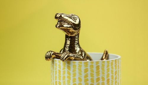 Free A shiny golden dinosaur figurine peeks out of a patterned cup against a yellow background. Stock Photo