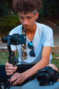A young man using a smartphone and gimbal for filming in a park setting, displaying modern photography techniques.