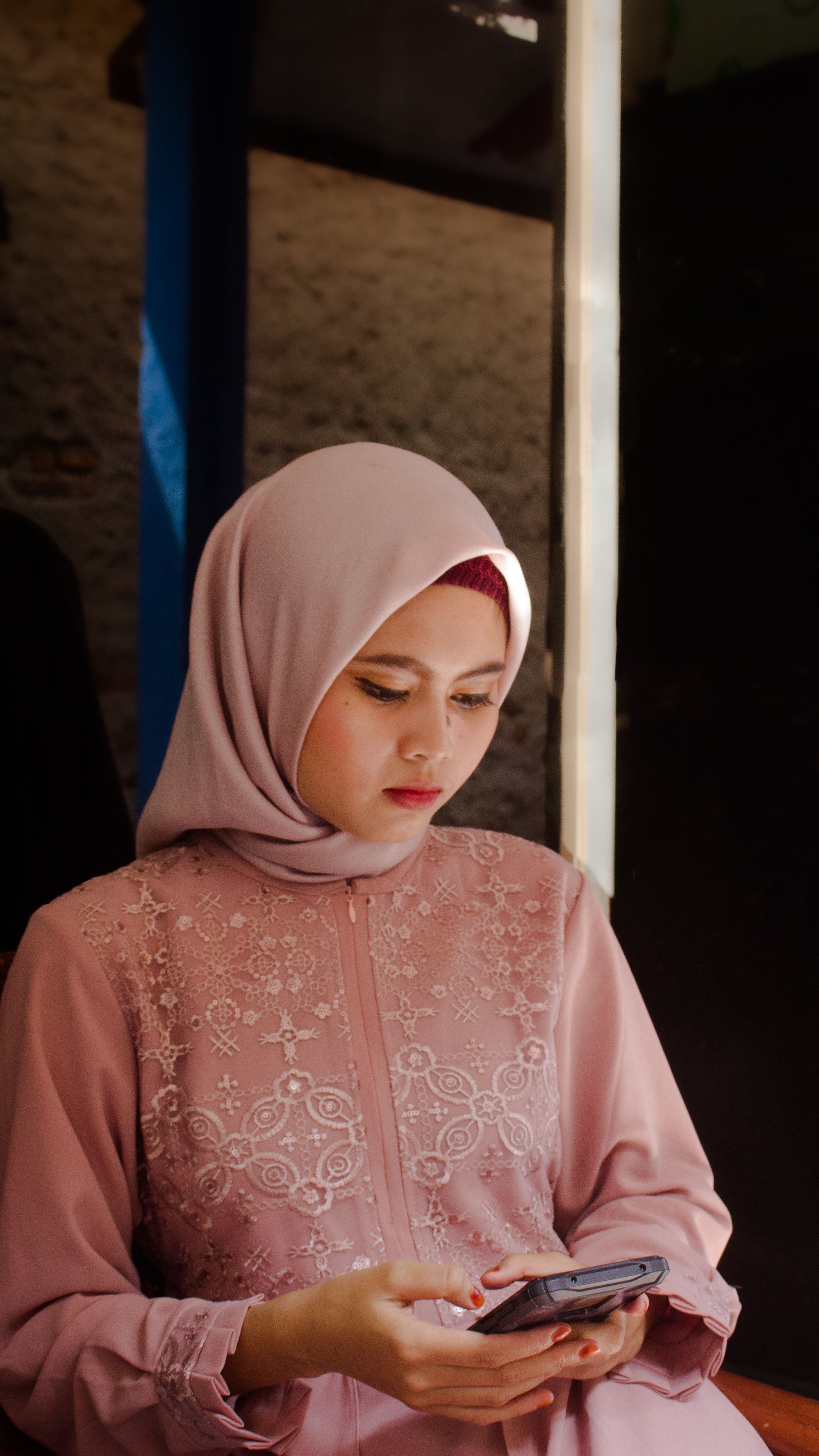 Young Muslim woman wearing a hijab, using a smartphone indoors in natural light.