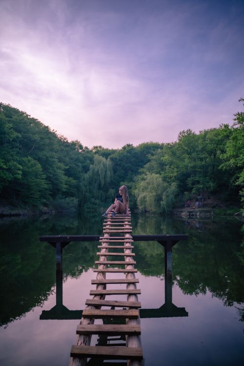 Free Tranquil scene of a girl sitting on a wooden bridge at dusk over a reflective river surrounded by lush greenery. Stock Photo