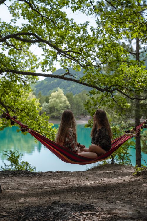 Free Two girls sit in a hammock, surrounded by lush greenery overlooking a tranquil lake. Stock Photo