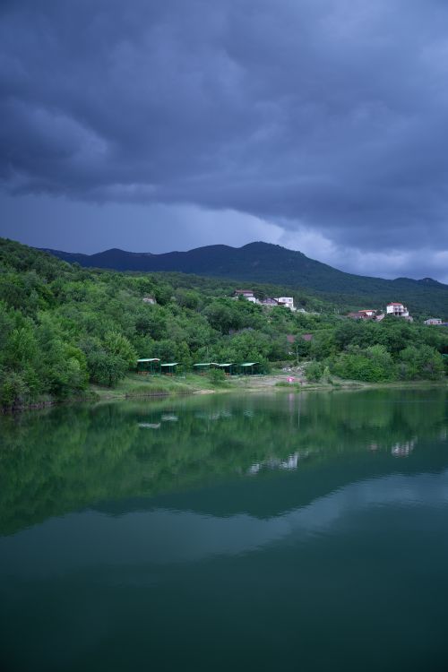 Free Calm lake surrounded by lush forest with dramatic overcast clouds and reflections. Stock Photo
