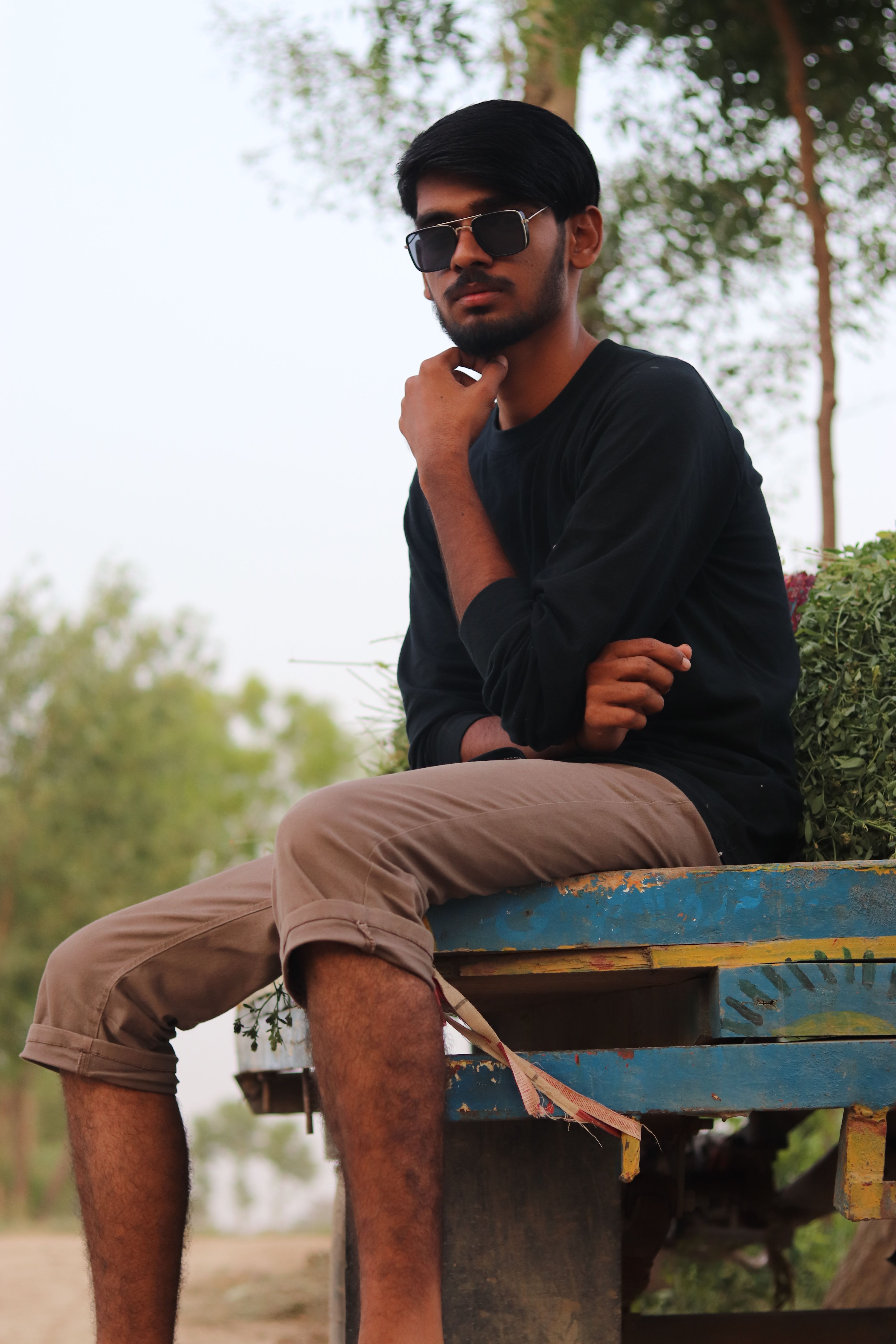 A fashionable man in sunglasses sits thoughtfully on a wooden bench outdoors.