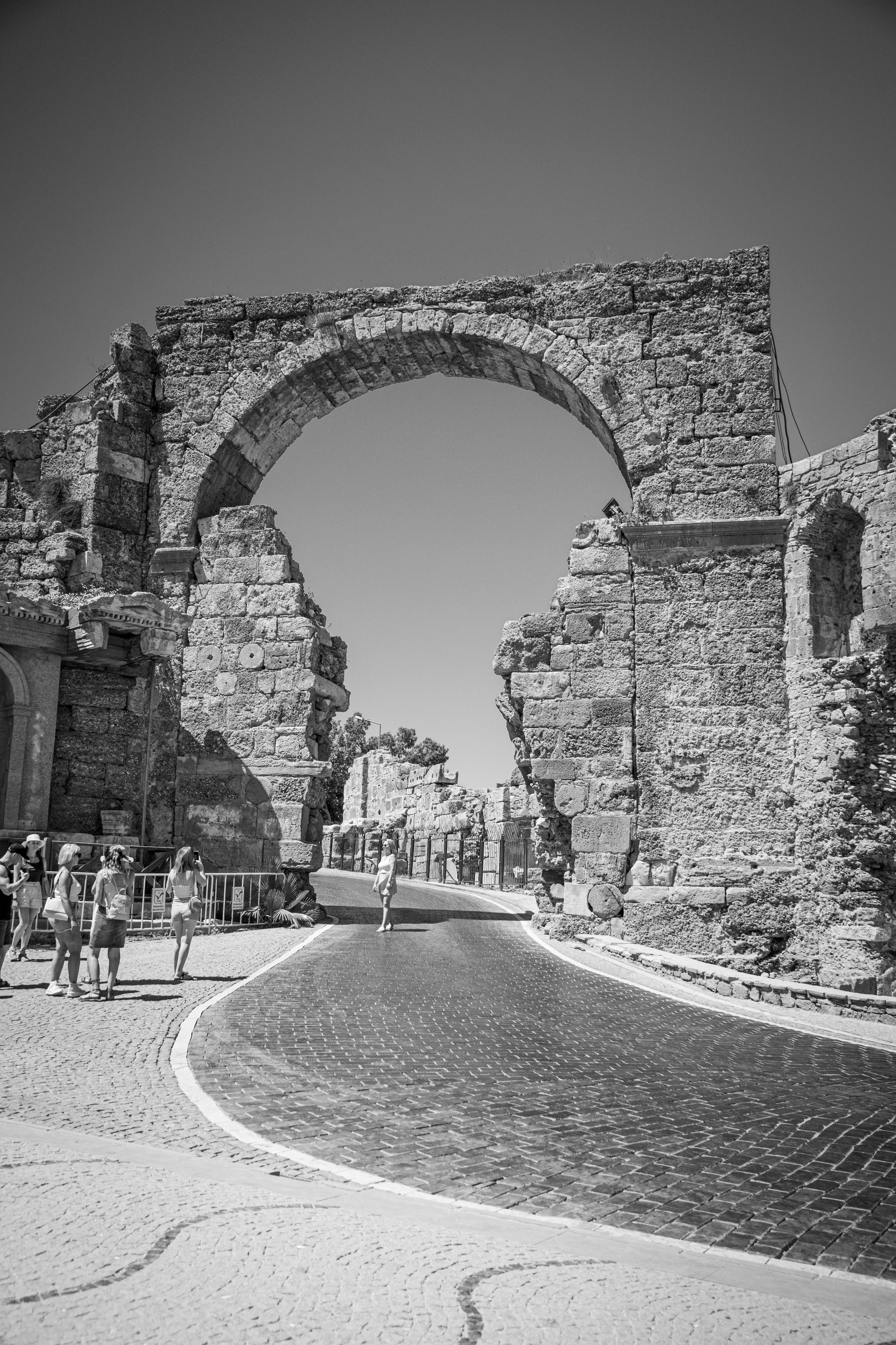 Black and white photo of a medieval stone archway over a cobblestone road with tourists.