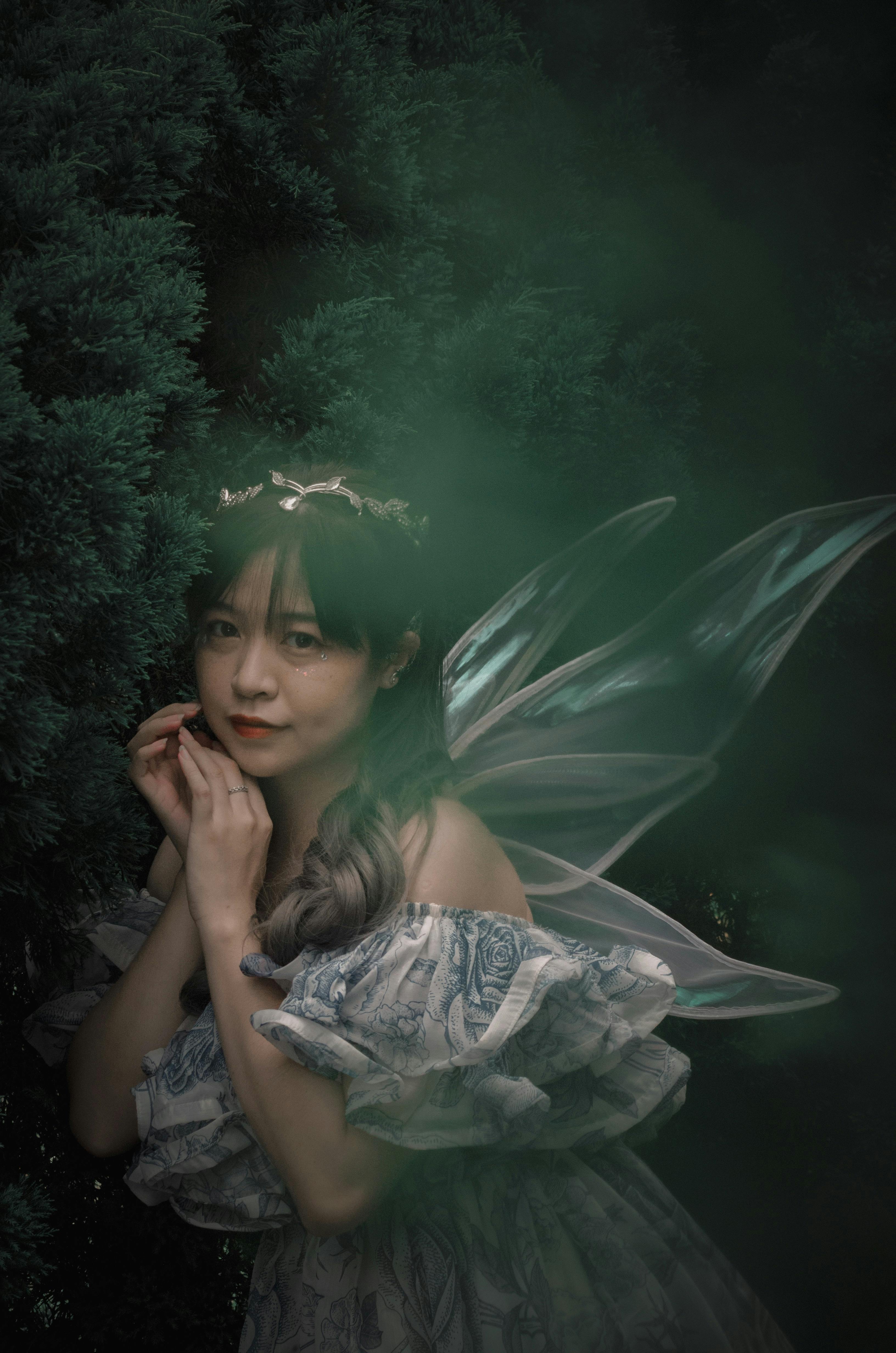 A young woman in fairy costume poses amidst lush greenery with ethereal wings.