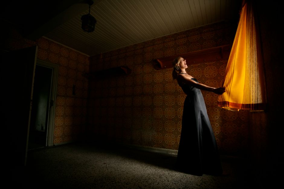 A woman stands by a glowing curtain in a vintage room, captured in dramatic lighting.