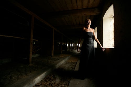 Женщина в черном платье без бретелек стоит у окна