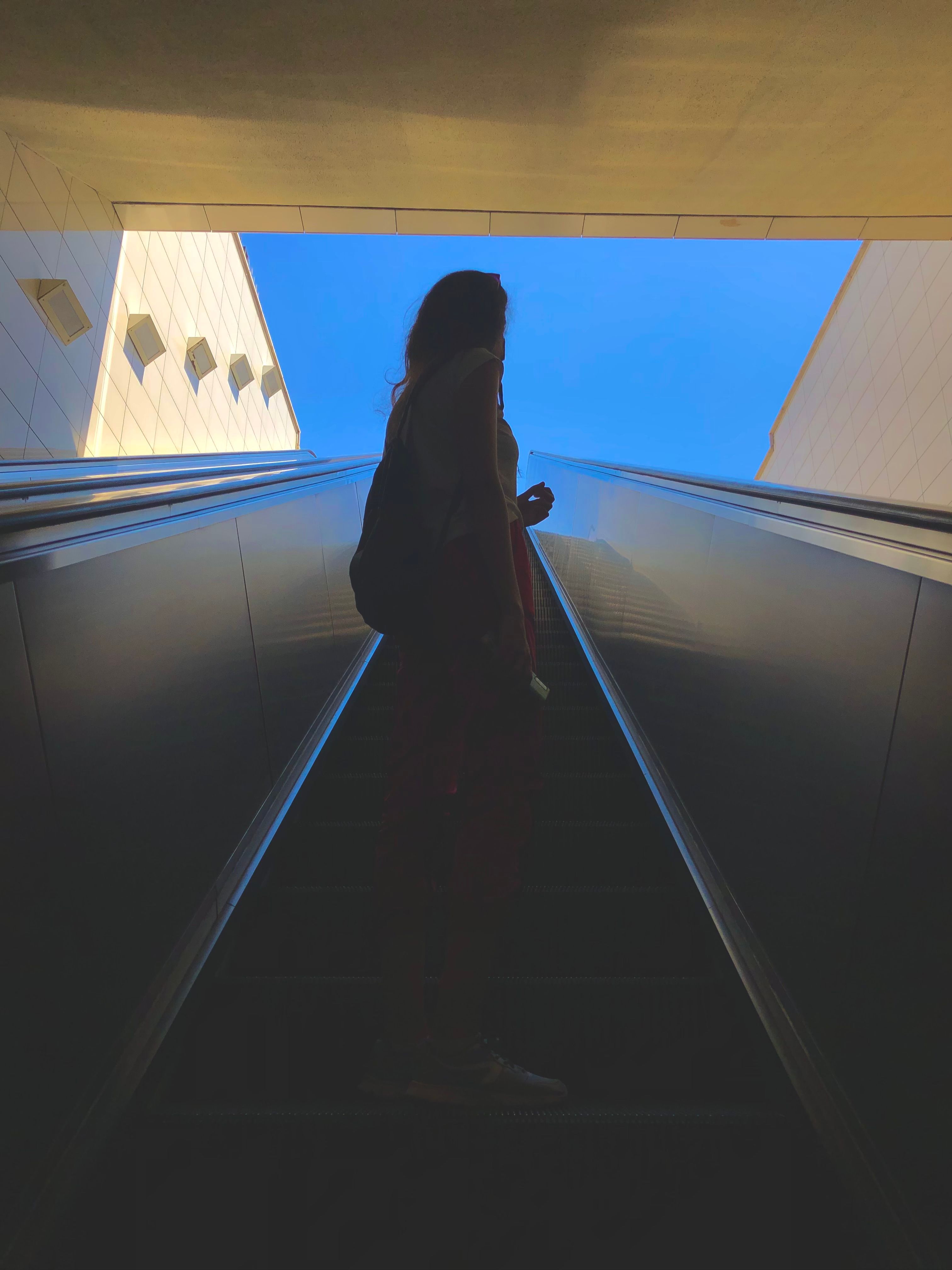 A dramatic silhouette of a woman standing on an escalator under a bright blue sky.