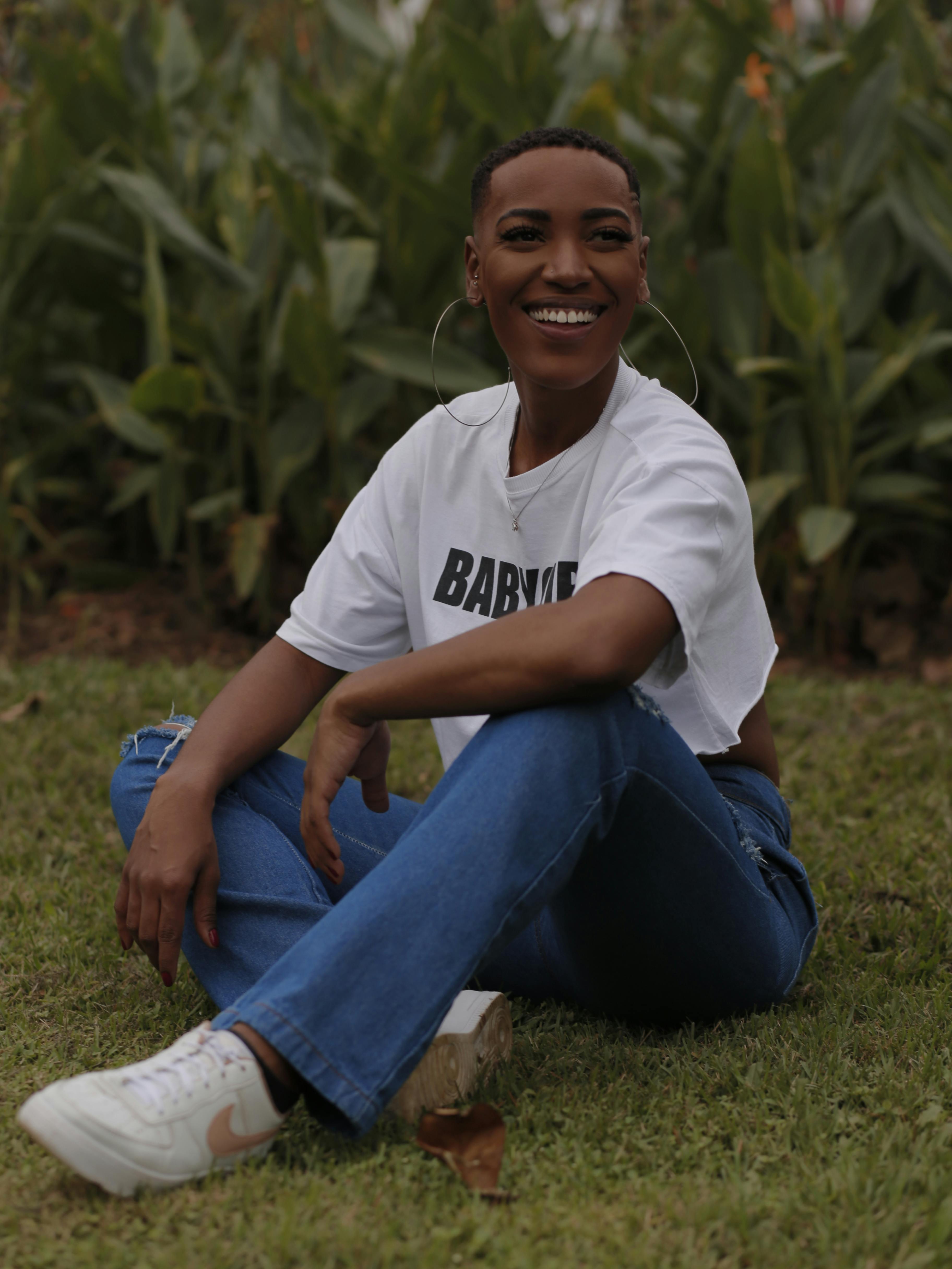 Young woman in casual wear sitting on grass, smiling joyfully amidst greenery.