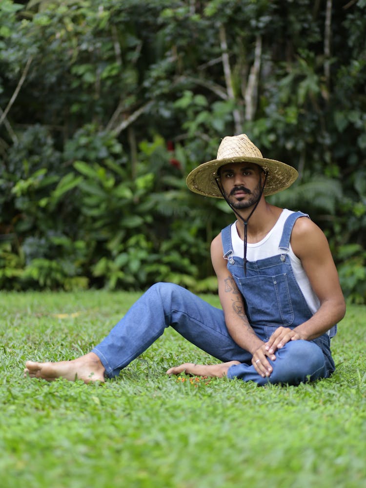 Man In Overalls And A Straw Hat Sitting On Grass Barefoot 