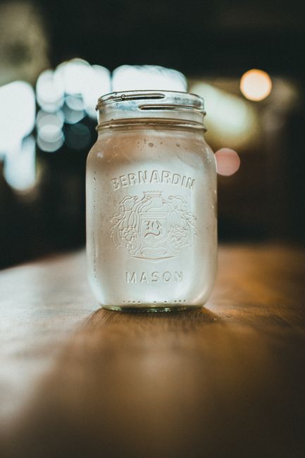 A mason jar filled with clear moonshine sitting on a wooden barrel, with a copper still visible in the blurred background.