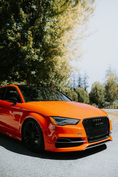 Free A sleek orange Audi parked under vibrant trees, showcasing elegance and luxury on a sunny road. Stock Photo