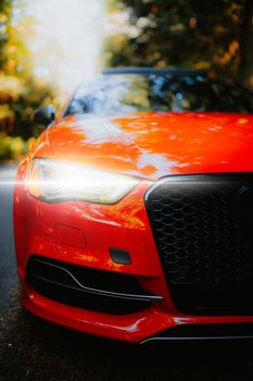 Close-up of a red car's front with illuminated headlights on a scenic road during the day.