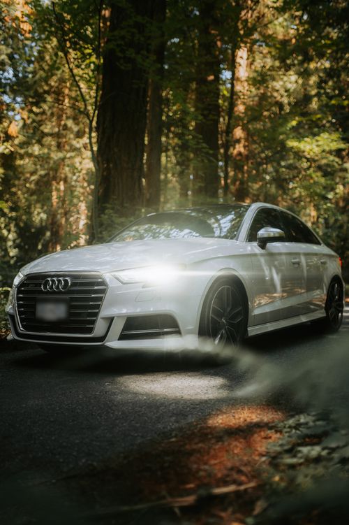 Free A sleek white luxury sedan with headlights on, parked on a forest road surrounded by towering trees. Stock Photo