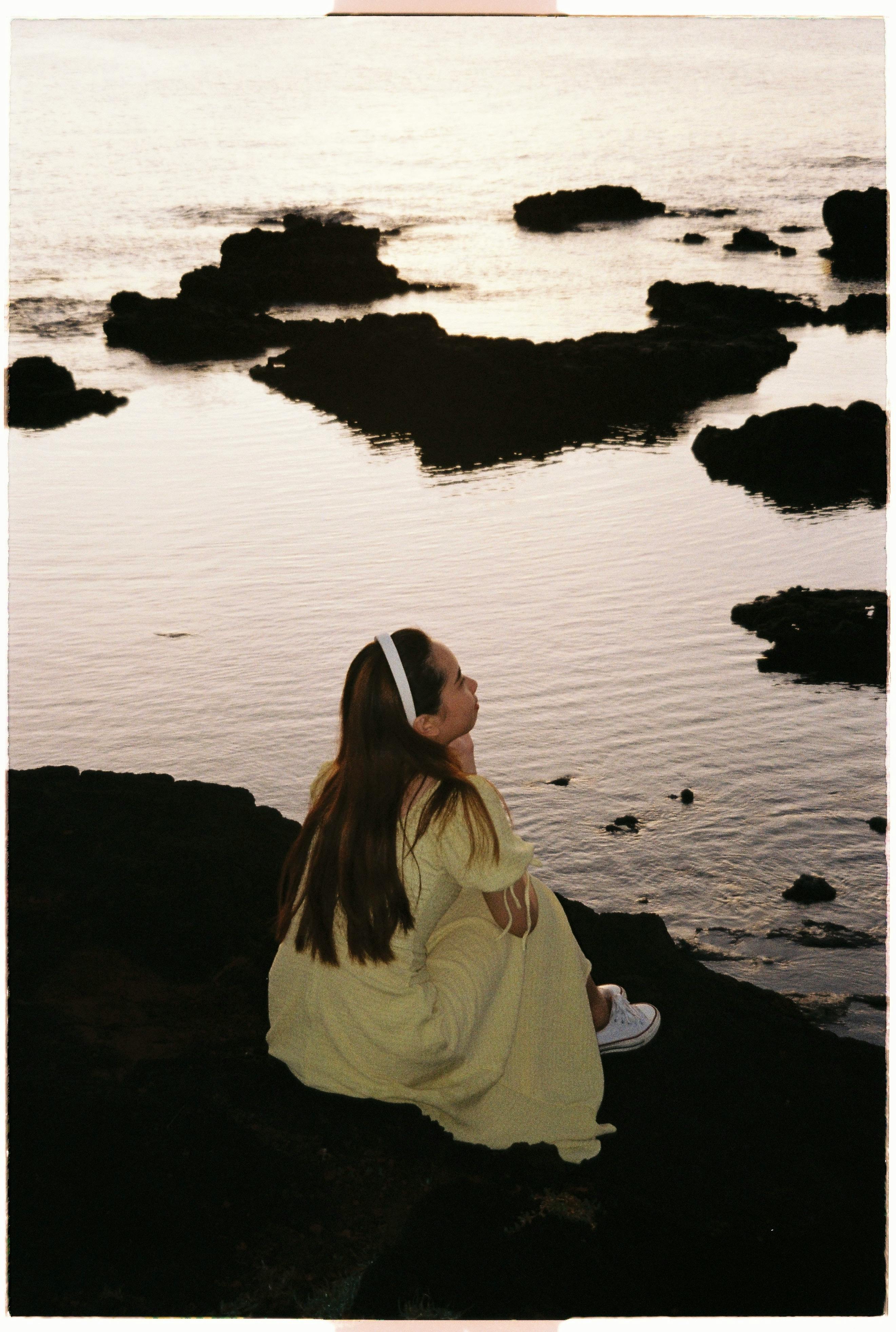 A woman in a yellow dress sitting by a rocky seaside at sunset, reflecting tranquility.