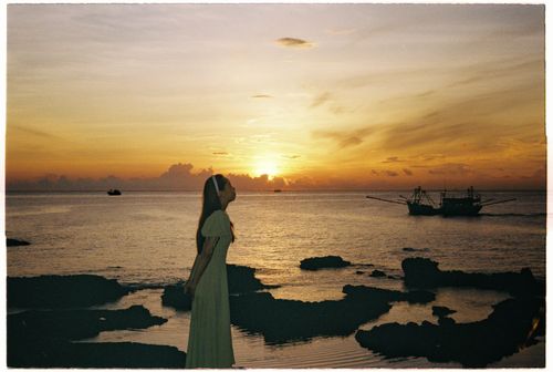 Free A serene seaside sunset with a silhouetted woman in a flowing dress by the rocks. Stock Photo