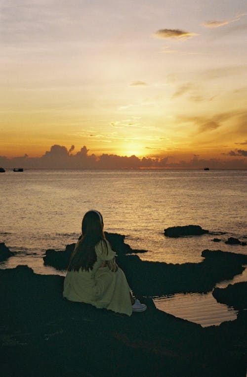 Woman in White Dress Sitting on Seashore during Sunset