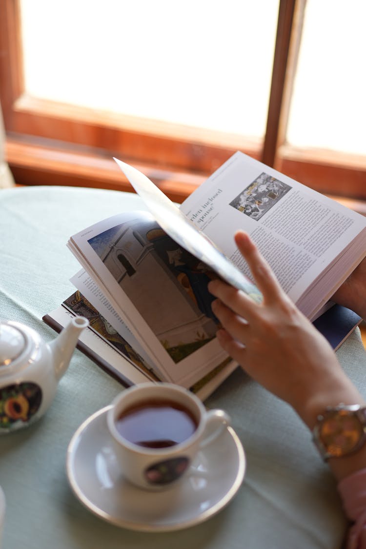 A Person Turning Pages Near The Table With Cup Of Tea