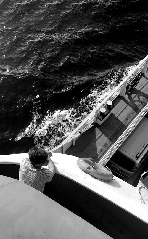 Grayscale Photo of a Man Standing on a Boat Sailing in the Sea
