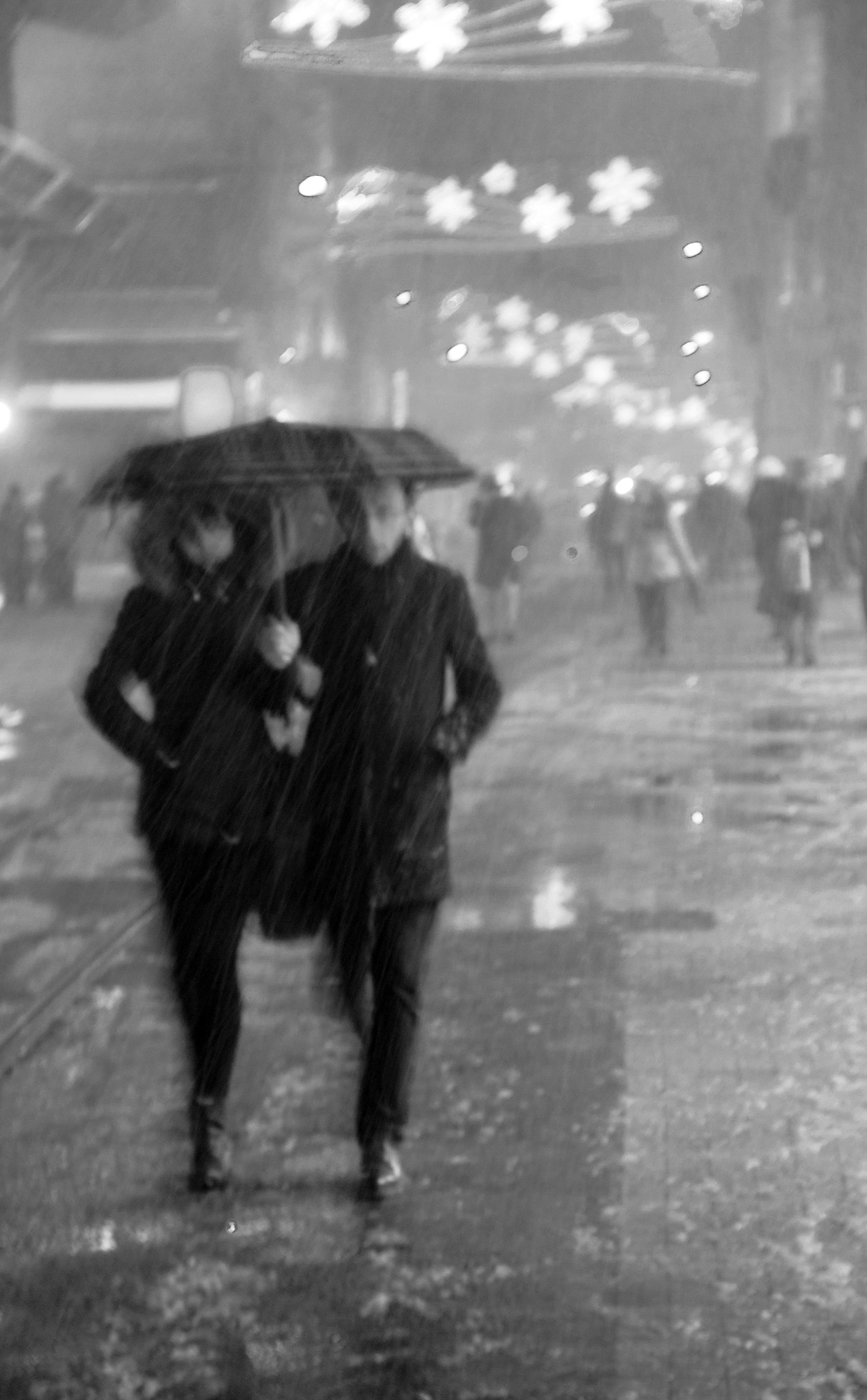 A couple walks under an umbrella during a night downpour on a city street, captured in black and white.