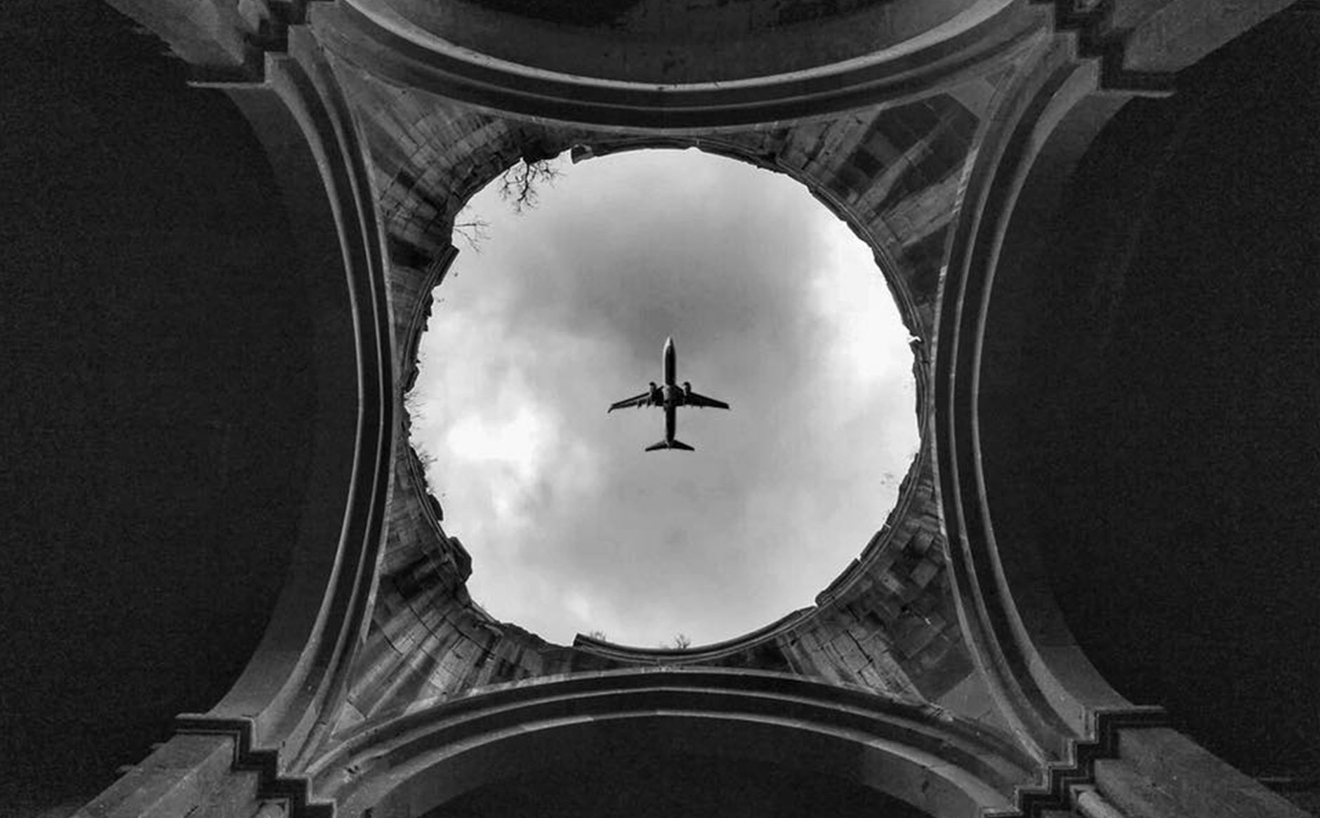 Airplane silhouetted against clouds seen through circular ruins, creating a dramatic perspective.