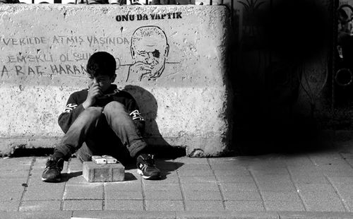 Free A child sits contemplatively on a sidewalk, leaning against a graffiti-covered wall in a city setting. Stock Photo