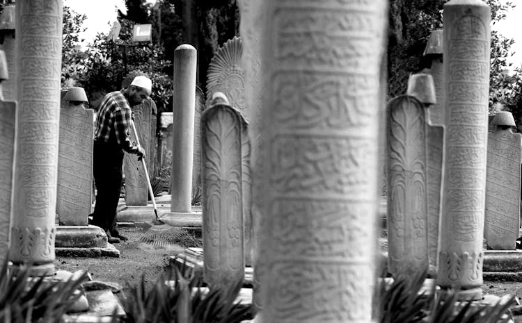 Black And White Photo Of A Man Raking The Ground Around Columns