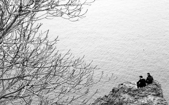 Black and white photo capturing two men sitting on a cliffside by the sea.