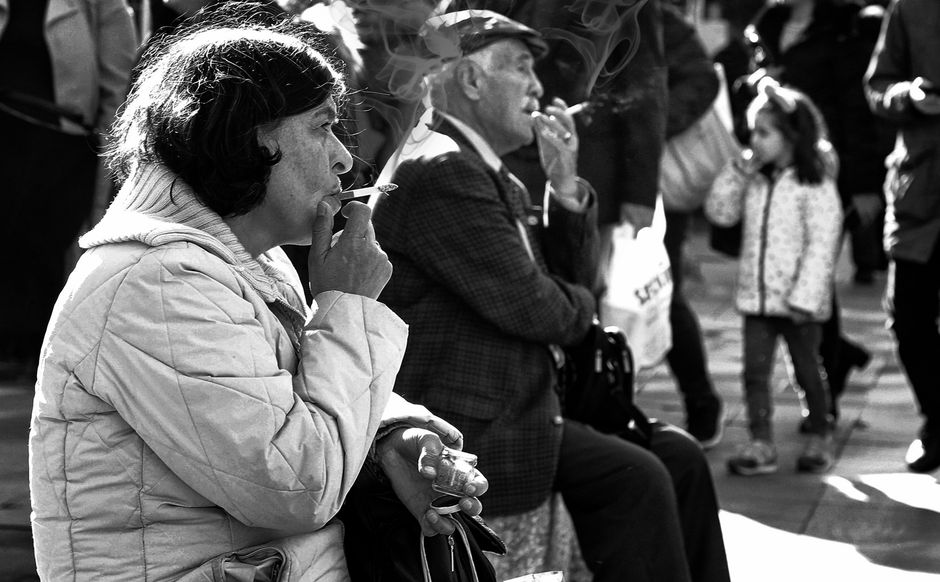 Hombre y mujer mayores fumando en una calle concurrida en imágenes en blanco y negro.