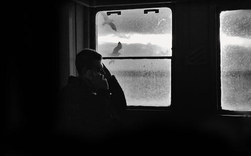 A Grayscale Photo of a Man Sitting Beside the Window