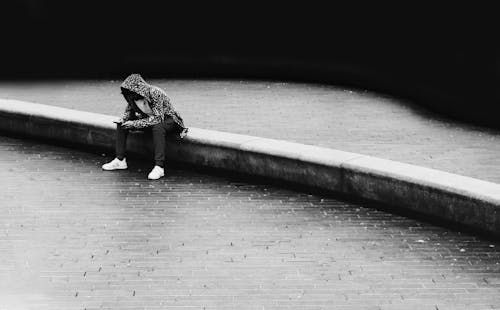A Grayscale Photo of a Person Sitting on the Gutter at the Street