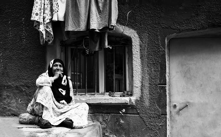 Old Woman In Hijab Sitting Outside Building On Street