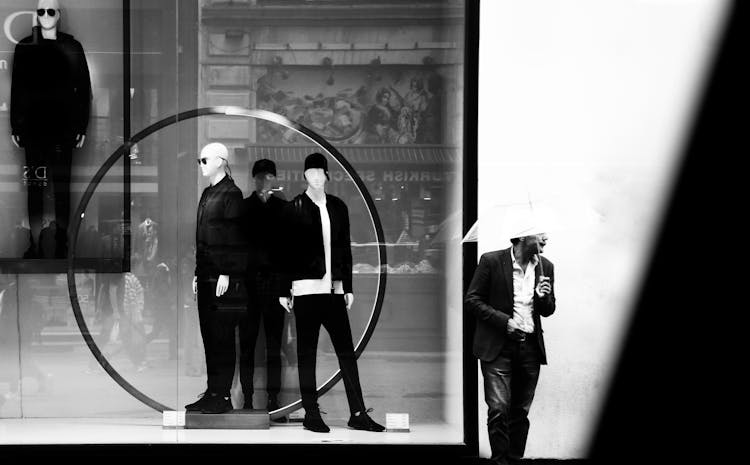 A Grayscale Photo Of A Man Walking On The Street Near The Store With Mannequins