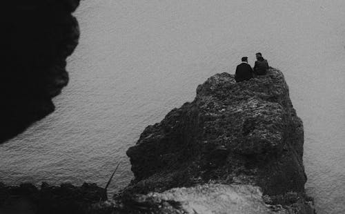 Grayscale Photo of People Sitting on a Cliff