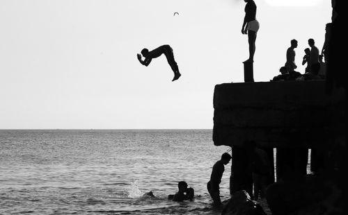 Free Silhouettes of people cliff jumping into the sea in a scenic black and white beach setting. Stock Photo