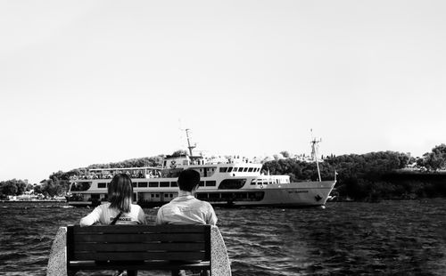 Free A couple sitting on a bench, gazing at a ferry passing by on the water. Stock Photo