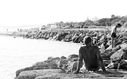 Grayscale Photo of Topless Man Sitting on Rock Near Body of Water
