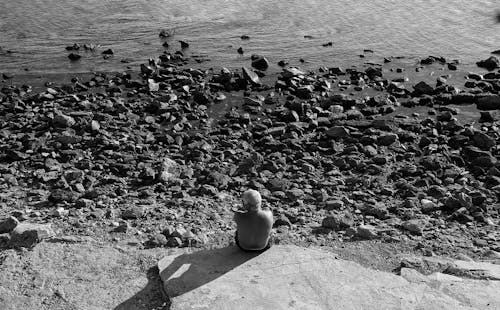 Free Grayscale Photo of a Man Sitting on a Rocky Seashore Stock Photo