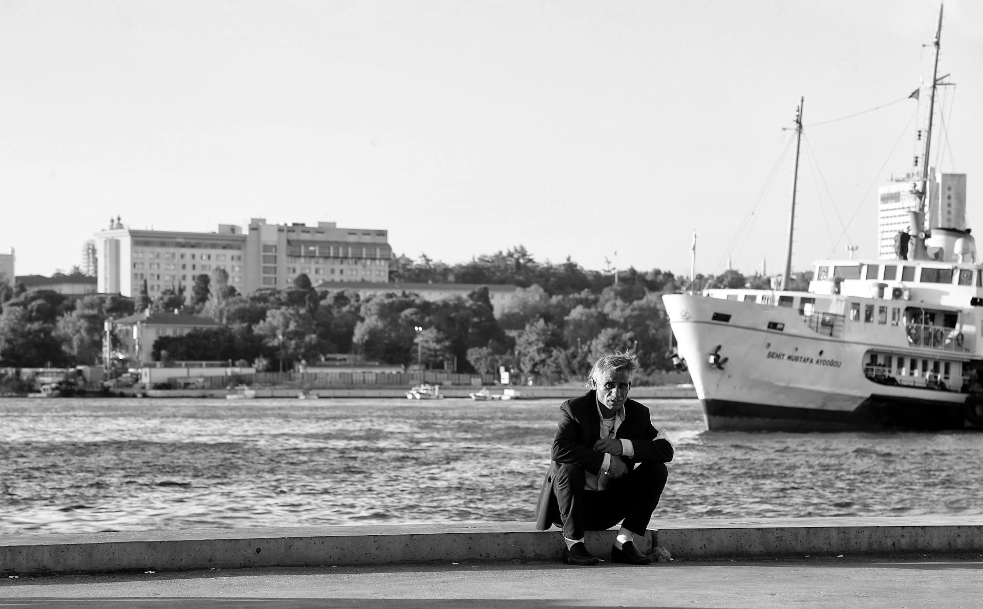 Elderly man sitting by the harbor with a large ship in the background.