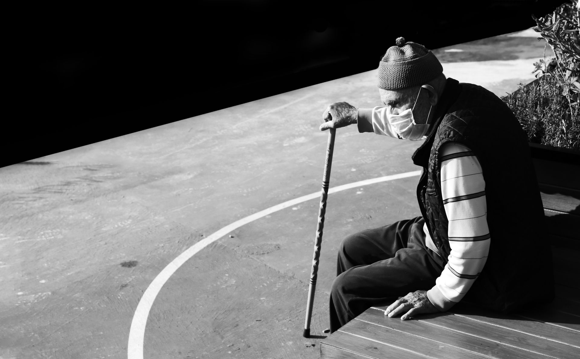 Elderly man wearing a face mask sitting outdoors with a walking stick.