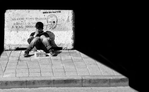 Free Black and white photo of a child in shadows sitting by a graffiti-covered concrete barrier. Stock Photo