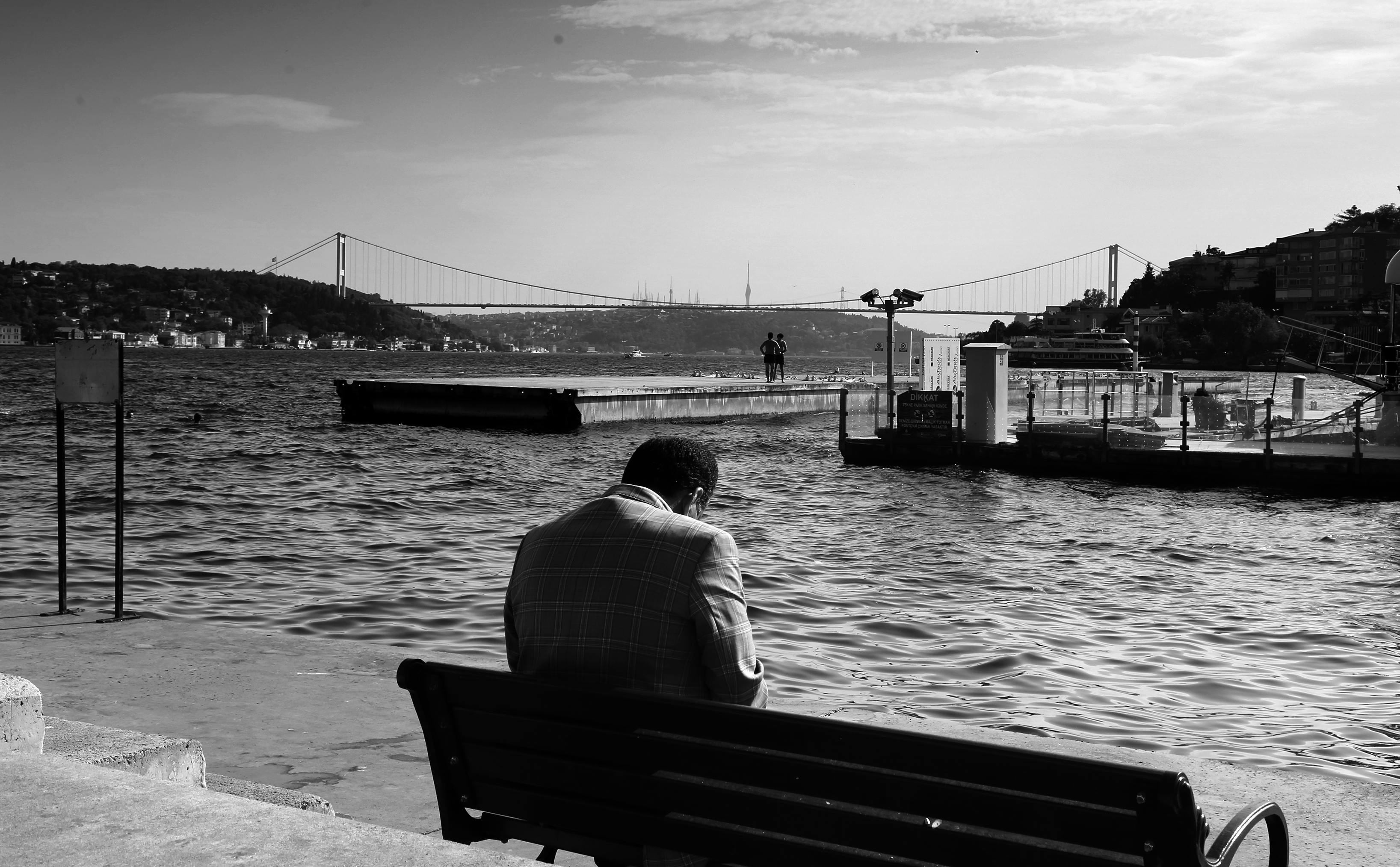 A man sitting on a bench by the Bosphorus, with Istanbul bridge in the background.